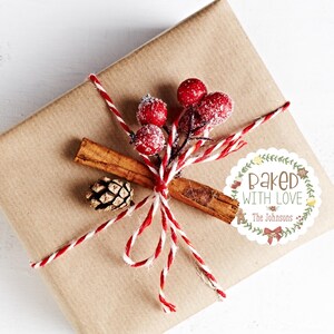 Op de afbeelding: Een cadeau verpakt in bruin papier, versierd met rood en wit touw, rode bessen, een kaneelstokje en een kleine dennenappel. Een witte sticker met de tekst "Baked with Love" en de achternaam is op het cadeau bevestigd.