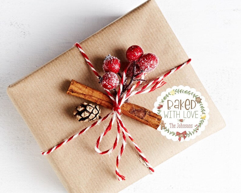 Op de afbeelding: Een cadeau verpakt in bruin papier, versierd met rood en wit touw, rode bessen, een kaneelstokje en een kleine dennenappel. Een witte sticker met de tekst "Baked with Love" en de achternaam is op het cadeau bevestigd.
