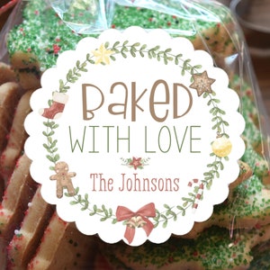 Op de afbeelding: Een doorzichtige cellofaan zak gevuld met kerstkoekjes, sommige met groene hagelslag. Een rond etiket leest "Baked With Love" met een krans en vakantie-illustraties. De tekst "The Johnsons" staat ook op het etiket.