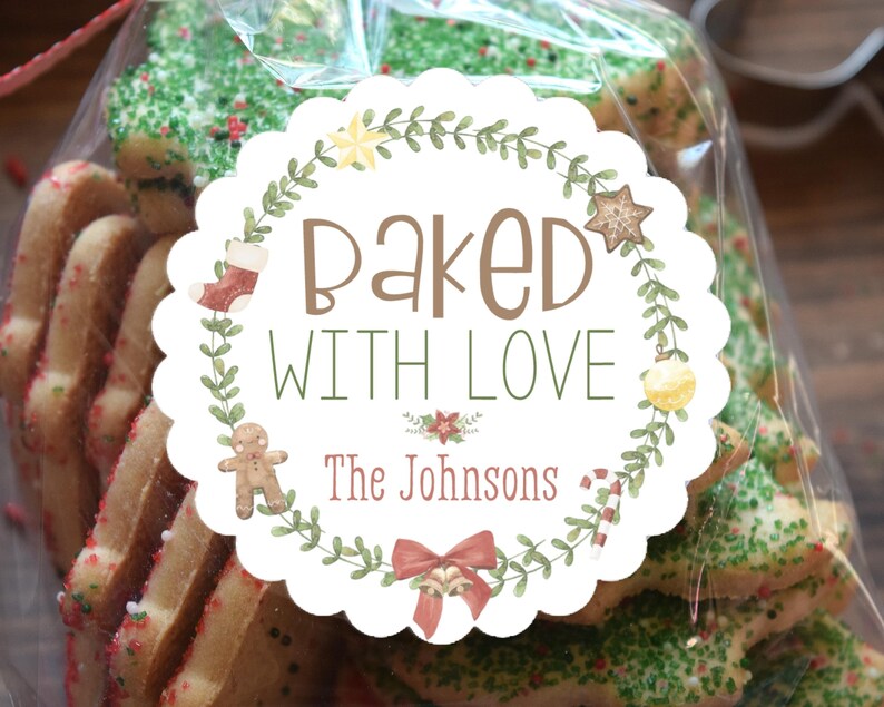 Op de afbeelding: Een doorzichtige cellofaan zak gevuld met kerstkoekjes, sommige met groene hagelslag. Een rond etiket leest "Baked With Love" met een krans en vakantie-illustraties. De tekst "The Johnsons" staat ook op het etiket.