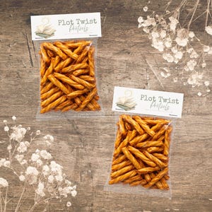 Può includere: Due sacchetti trasparenti pieni di pretzel dorati, ciascuno con un'etichetta bianca con la scritta "Plot Twist Pretzels". I sacchetti sono disposti su una superficie di legno rustico, con elementi floreali.