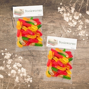 Puede incluir: Dos bolsas transparentes llenas de gusanos de goma de colores, rojos, amarillos y verdes, sobre una superficie de madera. Cada bolsa tiene una etiqueta que dice "Bookworms" con una ilustración de una pila de libros.