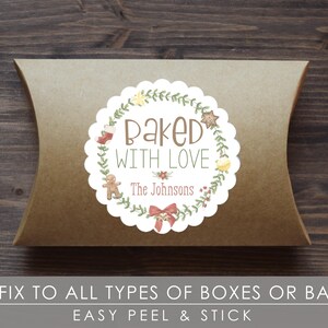 Op de afbeelding: Een bruine kussendoos met een witte sticker met geschulpte rand. De sticker leest "Baked With Love" en bevat een krans van groen, peperkoekmannetjes en strikillustraties. De tekst onderaan luidt "The Johnsons".