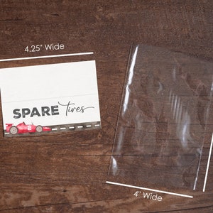Puede incluir: Una tarjeta blanca con el texto "SPARE Tires" y una ilustraci&oacute;n de coche de carreras rojo, 10,8 cm de ancho y 7,6 cm de alto. Una bolsa de pl&aacute;stico transparente, 10,2 cm de ancho y 15,2 cm de alto, est&aacute; al lado de la tarjeta. Los art&iacute;culos est&aacute;n sobre una superficie de madera.