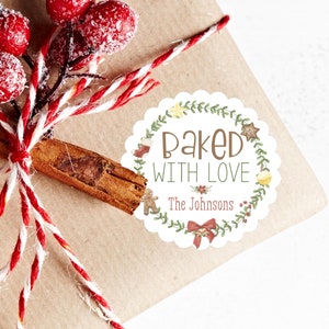 Op de afbeelding: Een ingepakt cadeau met rood-wit touw, een kaneelstokje en rode bessen. Het cadeau heeft een ronde sticker met de tekst "Baked with Love" en "The Johnsons" met feestelijke illustraties.