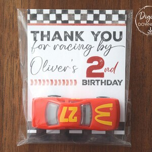 Może przedstawiać: Czerwony zabawkowy samochód wyścigowy z żółtymi akcentami i numerem 27 jest zapakowany z kartką z napisem "THANK YOU for racing by Oliver's 2nd BIRTHDAY". Karta ma wzór szachownicy na górze.