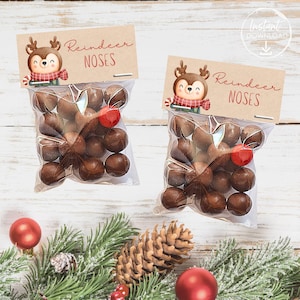 May include: Two clear cellophane bags filled with chocolate candies, labeled "Reindeer Noses." Each bag has a brown tag with a cartoon reindeer illustration and red text. Red ball ornaments, pine needles, and a pine cone are in the background.