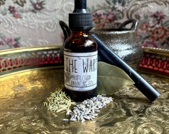 May include: A dark amber glass dropper bottle with a white label reading THE WARD PROTECTION ANOINTING OIL, accompanied by dried rosemary, lavender buds, a bay leaf, and a black wand, all resting on a gold-toned metal tray with ornate patterns.
