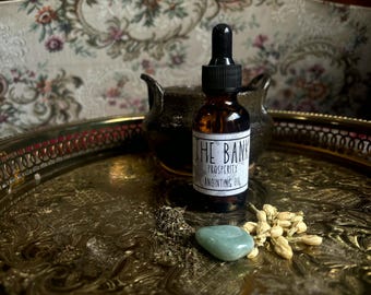 May include: A brown glass dropper bottle labeled THE BANK PROSPERITY ANOINTING OIL, positioned on a gold-toned metal tray with a small green polished stone, a pile of dried herbs, and a cluster of dried jasmine flowers nearby.