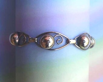 Może przedstawiać: Srebrna bransoletka z trzema sferycznymi elementami połączonymi ozdobnymi ogniwami. Centralna kula jest większa, z wirującym wzorem po obu stronach. Bransoletka ma polerowane wykończenie i odbija otoczenie.