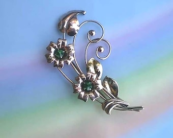 May include: A silver-toned metal brooch featuring two five-petaled flowers with small, round, vibrant green stones at their centers. The design includes curved metal stems, three smooth leaves, and two delicate wire swirls with small ball tips.