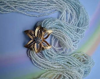 Może przedstawiać: Wielowarstwowy naszyjnik składający się z licznych jasnoniebieskich koralików typu seed beads zebranych razem. Złota metalowa broszka w kształcie kwiatu z sześcioma teksturowanymi płatkami i grupą przezroczystych cyrkonii w środku służy jako zapięcie.