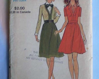 May include: A vintage Vogue sewing pattern envelope featuring illustrations of two outfits. One figure wears a long-sleeved beige top with a dark green skirt and belt. The other figure wears a short-sleeved red dress with a collared neckline.