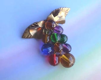 May include: A brooch featuring a cluster of multi-colored translucent glass beads shaped like grapes, suspended from a gold-toned metal leaf design. The beads are in shades of amber, purple, green, blue, and red, creating a vibrant, dangling effect.