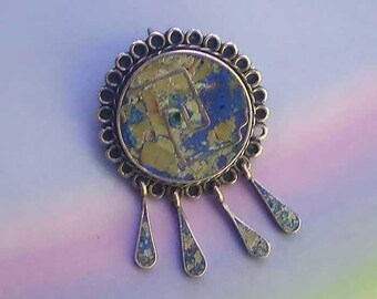 May include: A circular brooch featuring a central disc with a mottled blue and gold surface and a geometric line design. The disc is framed by a scalloped metal border and has four dangling, teardrop-shaped charms with matching blue and gold textures.