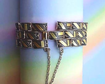 May include: A multi-row bracelet featuring rectangular black links with gold-toned diagonal stripes and etched patterns. The bracelet includes a secure clasp mechanism and a delicate gold-toned safety chain connecting the two ends.