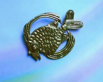 May include: A gold-toned metal brooch shaped like a fish. The fish features a textured, scale-like pattern on its body, a prominent circular eye, and elongated, curved fins that wrap around the body. The metal has a matte, slightly weathered finish.