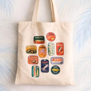 May include: A cream-colored canvas tote bag featuring a colorful graphic print of various canned seafood labels, including sardines, octopus, mussels, smoked oysters, squid, smoked salmon, clams, mackerel, anchovies, and tuna, arranged in a scattered pattern.