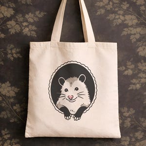 May include: Cream-colored tote bag featuring a cartoon opossum illustration within a black scalloped circle. The opossum has gray fur, a pink nose, and black paws. The bag hangs from a gold hook against a dark floral patterned background.
