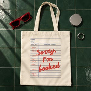 May include: A cream-colored canvas tote bag featuring a blue library checkout card graphic with the red text "Sorry, I'm booked" printed in the center. The bag has two matching fabric handles and is displayed alongside red sunglasses and a silver mirror.