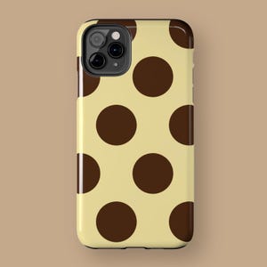 May include: A phone case featuring a pattern of large, dark brown polka dots on a light yellow background. The case has a glossy finish and a camera cutout.