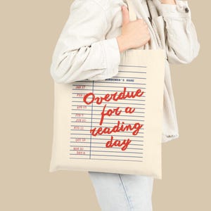 Puede incluir: Un bolso de tela color crema con un dise&ntilde;o de tarjeta de pr&eacute;stamo de biblioteca vintage. El estampado incluye l&iacute;neas horizontales azules, fechas, el texto "BORROWER'S NAME" en azul y la frase en cursiva roja "Overdue for a reading day" centrada.