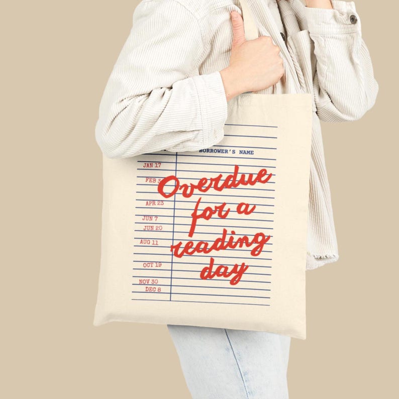 Puede incluir: Un bolso de tela color crema con un dise&ntilde;o de tarjeta de pr&eacute;stamo de biblioteca vintage. El estampado incluye l&iacute;neas horizontales azules, fechas, el texto "BORROWER'S NAME" en azul y la frase en cursiva roja "Overdue for a reading day" centrada.