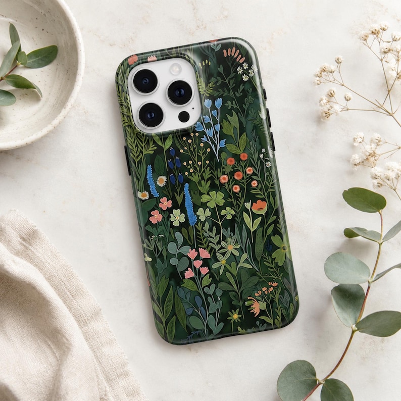 Pu&ograve; includere: Una cover per smartphone verde scuro con un'illustrazione botanica dettagliata di vari fiori selvatici e fogliame nei toni del rosa, arancione, blu e bianco. La cover presenta un ritaglio per un sistema a tripla fotocamera in alto a sinistra.