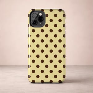 May include: A phone case with a repeating pattern of dark brown polka dots on a light yellow background. The case has a black camera module and a glossy finish. The design is simple and classic.