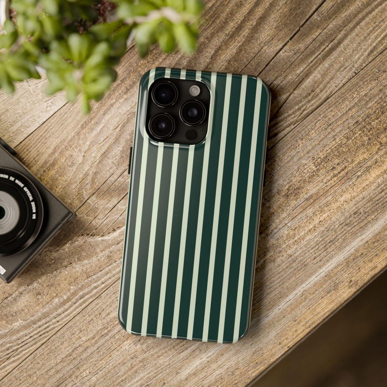 Może przedstawiać: Etui na smartfon z pionowym wzorem w paski w kolorze ciemnozielonym i jasnozielonym. Etui leży na drewnianej powierzchni obok czarnego aparatu i zielonej rośliny.