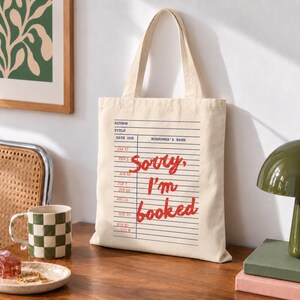 May include: A cream-colored canvas tote bag featuring a blue library checkout card graphic with horizontal lines and the red text "Sorry, I'm booked" printed in the center. The bag has two matching fabric handles and a rectangular vertical shape.