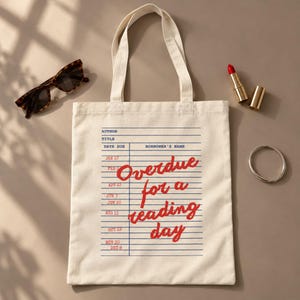 Puede incluir: Una bolsa de tela color crema con un gr&aacute;fico de tarjeta de biblioteca azul y el texto rojo "Overdue for a reading day" impreso en el frente. La bolsa tiene dos asas de tela a juego y una forma rectangular con l&iacute;neas horizontales azules.