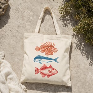 May include: A cream-colored canvas tote bag featuring a vertical graphic print of three fish. The top fish is orange, the middle fish is blue, and the bottom fish is red. The bag has two matching fabric handles and a simple rectangular shape.