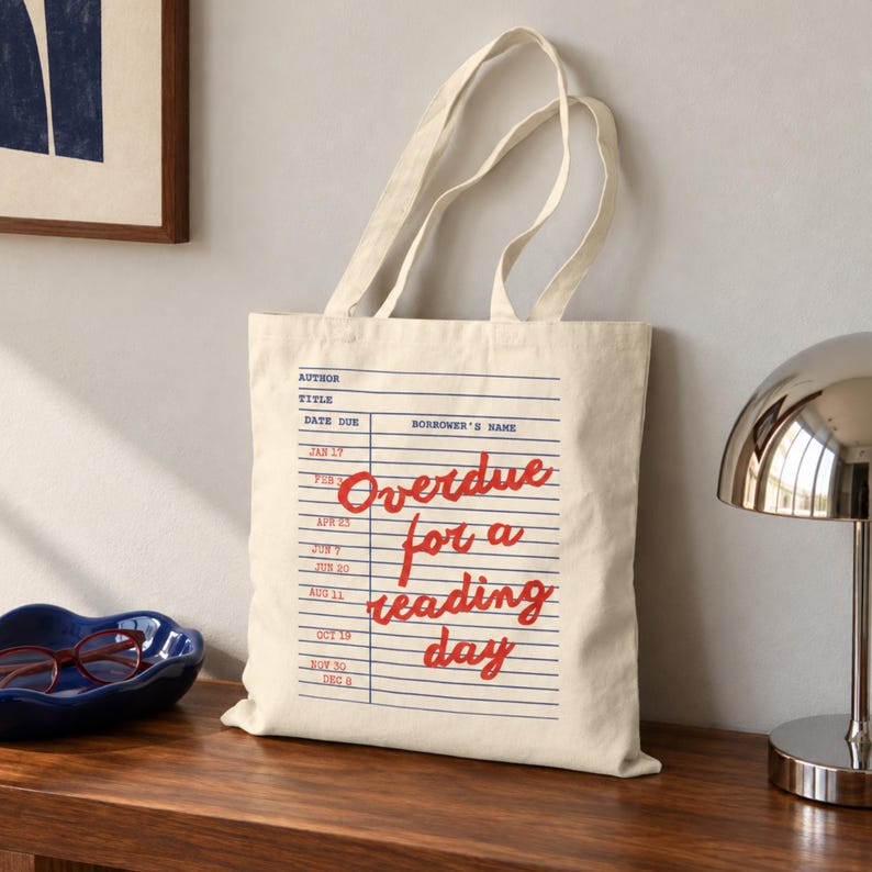 Puede incluir: Un bolso de tela color crema con un dise&ntilde;o impreso en azul de una ficha de biblioteca con l&iacute;neas y columnas horizontales. El texto en cursiva roja dice Overdue for a reading day, superpuesto sobre la cuadr&iacute;cula de la ficha bibliotecaria.