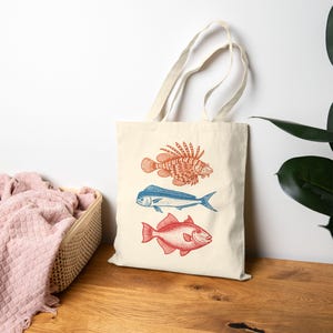 May include: A cream-colored canvas tote bag featuring a vertical graphic print of three fish. The top fish is orange, the middle fish is blue, and the bottom fish is red. The bag has two long fabric handles and a simple rectangular shape.