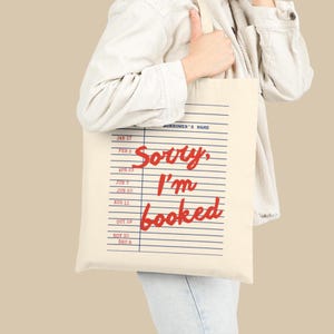 May include: A cream-colored canvas tote bag featuring a printed design that mimics a library book checkout card. The graphic includes blue horizontal lines, dates, and the red cursive text that reads, Sorry, I'm booked, with Borrower's Name at the top.
