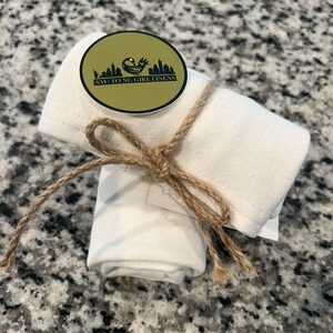 Può includere: Due asciugamani da cucina in cotone bianco arrotolati e legati con spago. Un adesivo dorato con testo nero recita "NYC to NC Girl Linens" e una piccola grafica di un uccello.