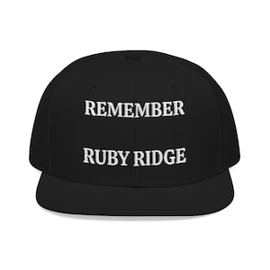 May include: Black baseball cap with a flat brim. The cap features the white text "REMEMBER" above "RUBY RIDGE". The hat is a classic style, suitable for casual wear. The text is centered on the front of the cap.