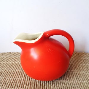 May include: A bright red ceramic pitcher with a rounded, spherical body and a curved handle. The interior of the pitcher and the rim are finished in a contrasting off-white color, showing minor wear along the top edge of the spout.