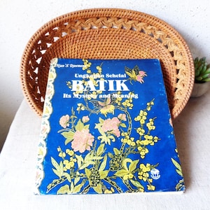 May include: A blue book titled "Batik: Its Mystery and Meaning" with floral and butterfly illustrations. The book is set against a woven wicker tray.
