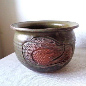 May include: A handcrafted ceramic pot with a dark green exterior and a reddish-brown carved design. The pot has a rounded shape with a wide opening and a slightly flared rim. The carved design appears to be a stylized bird or leaf.