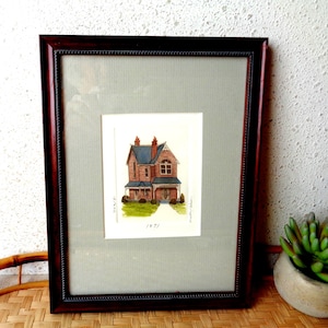 May include: Framed watercolor painting of a brick house with a blue roof and a porch. The artwork is set against a gray mat and framed in a dark wood frame. The painting is signed and dated 1971.