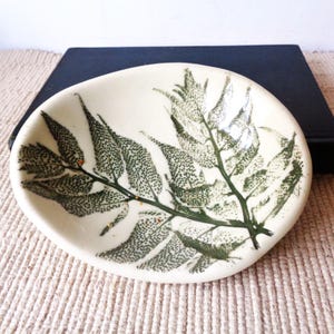 May include: An oval-shaped ceramic dish with a smooth, off-white glazed surface. The interior features a detailed, dark green botanical print of a fern frond, showcasing intricate leaf textures and a central stem extending across the basin.