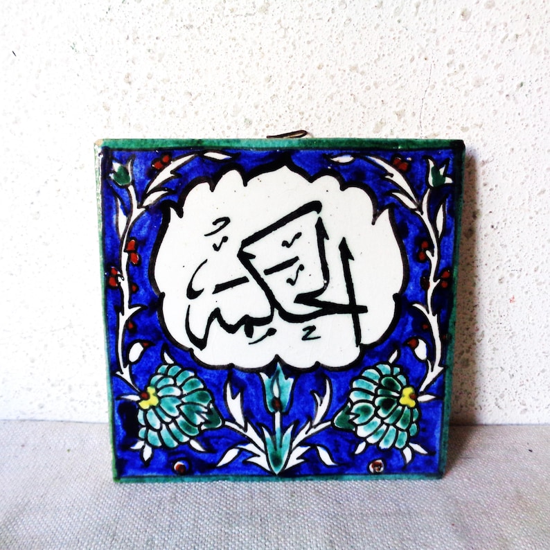 May include: Square ceramic tile with Arabic calligraphy in black on a white background. The tile is framed by a blue border with floral and leaf designs in green, white, and red. The tile has a green edge.