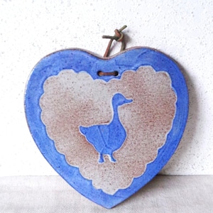 May include: Heart-shaped ceramic wall hanging with a blue border and a central design featuring a blue goose. The interior has a textured, off-white and brown pattern. A leather cord is attached for hanging.