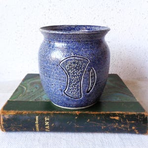 May include: A blue ceramic jar with a textured surface and a decorative design. The jar features a carved pattern on the front and is set on a vintage book. The jar's color is a speckled blue, and the design is a lighter shade of blue.
