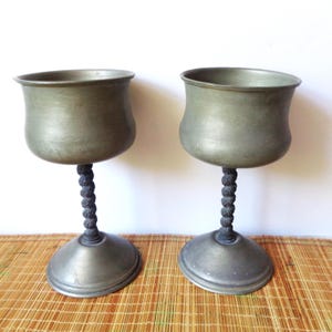 May include: Two antique pewter goblets with a dark, twisted stem and a wide, flared cup. The goblets have a classic, vintage design, perfect for display or use. The base is round and sturdy, and the overall color is a muted gray.