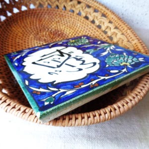May include: Square ceramic tile with a blue and white floral design. The tile has a white center with black calligraphy. The edges are green. The tile is resting in a woven basket.