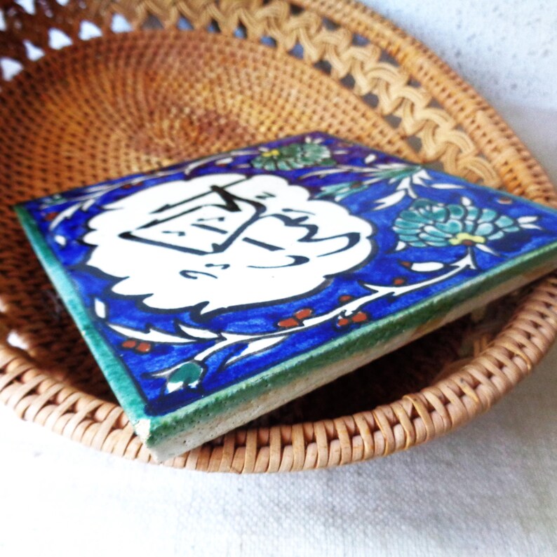 May include: Square ceramic tile with a blue and white floral design. The tile has a white center with black calligraphy. The edges are green. The tile is resting in a woven basket.