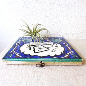May include: A square, ceramic tile with a blue and white floral design, featuring a central white cloud-shaped element with black calligraphy. A small air plant sits atop the tile. The tile has a green border and a metal clasp.